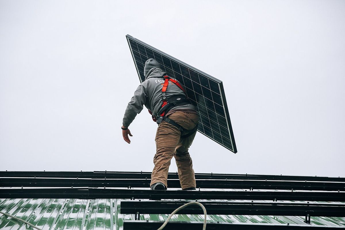 Técnico de costas carregando uma placa solar sobre um telhado